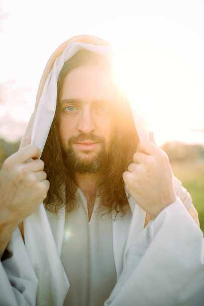 Portrait of Jesus Christ standing in meadow clothed in his traditional white robe against sunset background. - Photo, Image