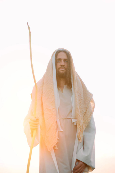 Jeesus Kristus puinen sauva seisoo hänen perinteisessä valkoisessa kaapussaan taivasta ja auringonlaskun taustaa vasten. - Valokuva, kuva