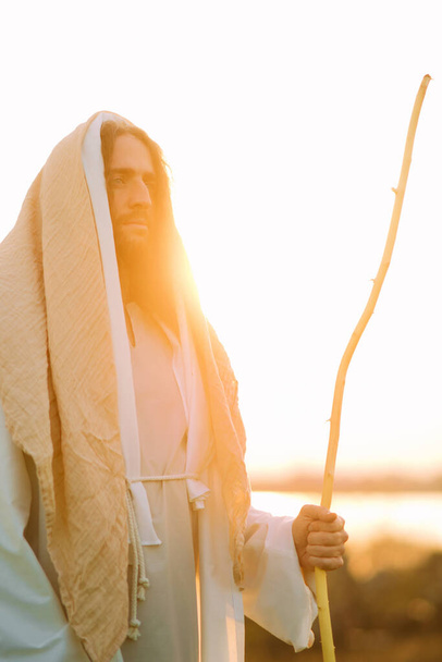 Jezus Chrystus z drewnianą laską stoi na łące ubrany w tradycyjną białą szatę na tle rzeki i zachodu słońca. - Zdjęcie, obraz