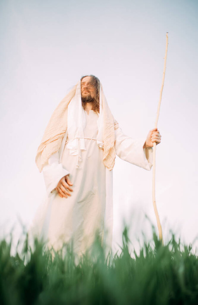 Jezus Chrystus z drewnianą laską stoi na łące ubrany w tradycyjną białą szatę na tle nieba. - Zdjęcie, obraz