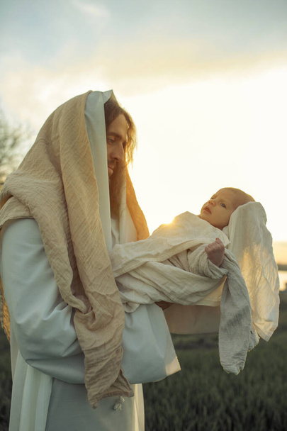 Святой Иосиф держит младенца Иисуса на руках и смотрит на него с любовью на лугу и фоне неба. - Фото, изображение