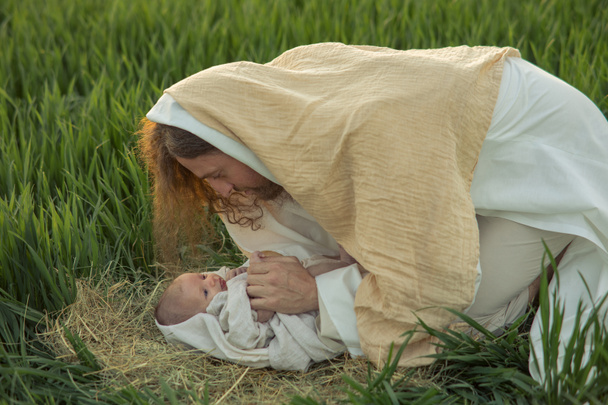 Святой Иосиф склоняется над младенцем Иисусом лежащим на траве и смотрит на него с любовью. - Фото, изображение