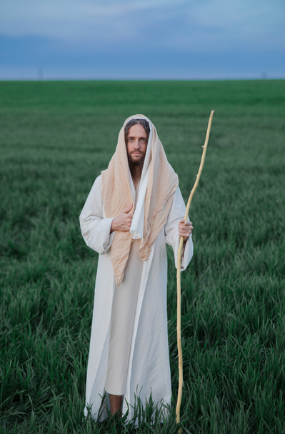 Jesus Christus mit Holzstab geht in seinem traditionellen weißen Gewand über die Wiese. - Foto, Bild