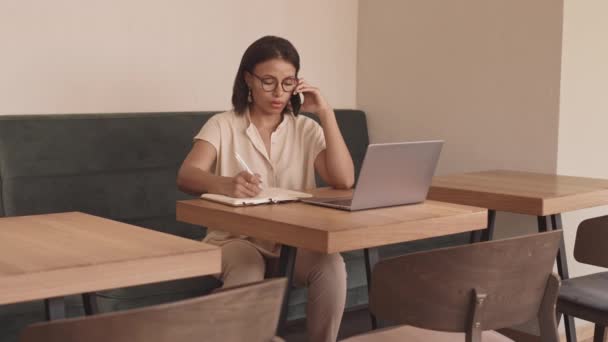 Longue moyenne de femme mixte moderne assise à table dans un restaurant, ayant un appel d'affaires sur un téléphone mobile, utilisant un ordinateur portable, écrivant dans un carnet - Séquence, vidéo