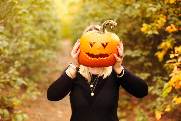 Close-up foto van jonge blonde vrouw houden eng vervaardigde pompoen in de voorkant van haar gezicht, geïsoleerd over herfst bos achtergrond. Herfstvakantie, Halloween decoraties en mensen concept - Foto, afbeelding