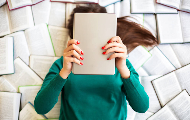 Uitzicht van boven van brunette vrouw in groene trui ligt op een groot aantal open boeken, terwijl het houden van digitale tablet in handen voor haar gezicht. Technologie, onderwijs en communicatie - Foto, afbeelding