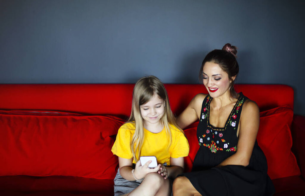 Happy smiling mother and daughter sitting on sofa in living room while using smartphone, cheerful mom and little child girl playing making photo on mobile, mommy and kid having fun with phone at home - Foto, imagen
