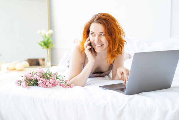 attraente zenzero donna mezza età in pigiama sdraiato a letto con telefono e laptop - Foto, immagini