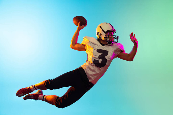 Portrait of American football player catching ball in jump isolated on blue studio background in neon light. - Фото, зображення