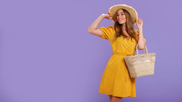 Beautiful smiling young woman in yellow dress, summer straw hat, bag isolated on pastel violet background.  - Fotografie, Obrázek
