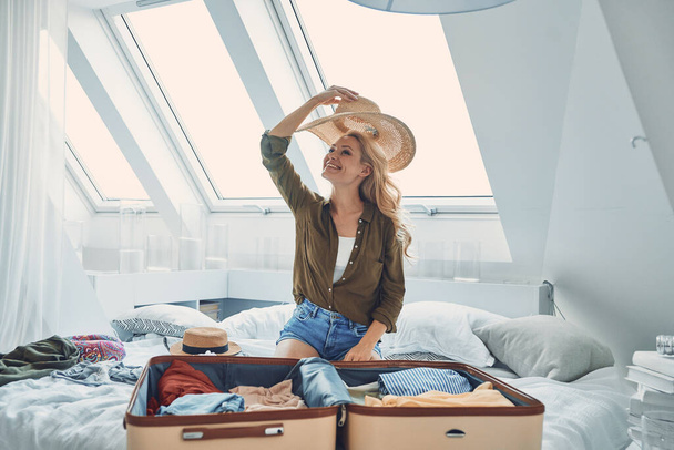 Beautiful young smiling woman packing luggage while sitting in bed at home - Photo, Image