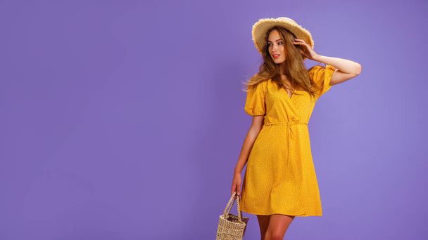 Beautiful smiling young woman in yellow dress, summer straw hat, bag isolated on pastel violet background.  - Φωτογραφία, εικόνα