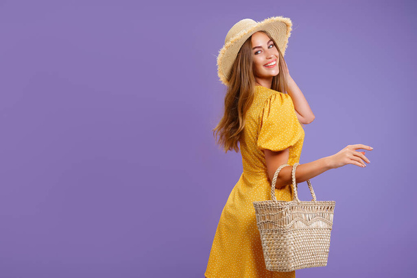Beautiful smiling young woman in yellow dress, summer straw hat, bag isolated on pastel violet background.  - Photo, image