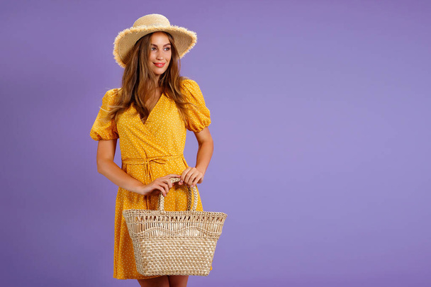 Piękna uśmiechnięta młoda kobieta w żółtej sukience, letni słomkowy kapelusz, torba odizolowana na pastelowym fioletowym tle.  - Zdjęcie, obraz