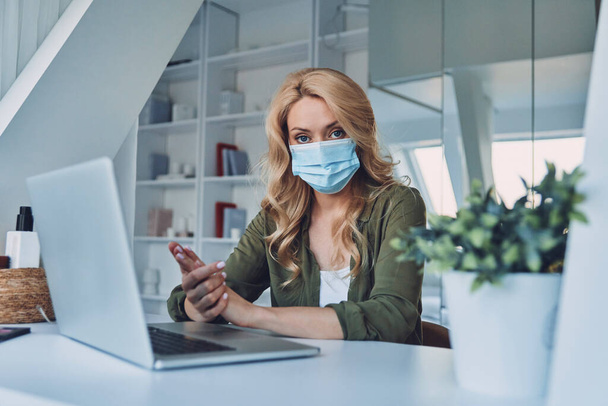 Attractive young woman in protective face mask sitting at the desk in office - Foto, Imagem