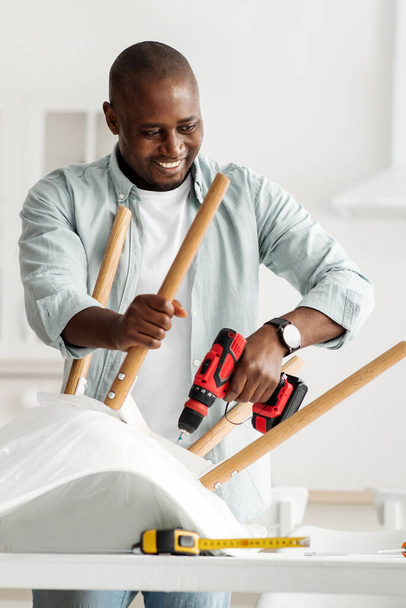 Kézi munka otthon. Boldog millenniumi fekete ember összeszerelése bútorok elektromos csavarhúzó a konyha belsejében, termés - Fotó, kép