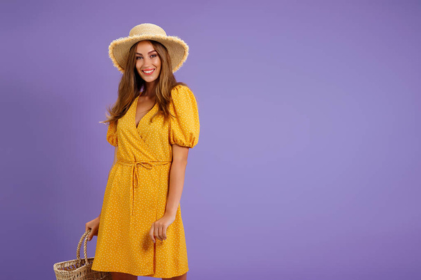 Beautiful smiling young woman in yellow dress, summer straw hat, bag isolated on pastel violet background.  - Zdjęcie, obraz