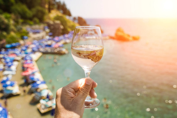 Um copo de vinho na mão. Copo com vinho branco fresco jovem no fundo da praia do Mediterrâneo e do mar em uma cidade turística no verão sob a luz do sol. Verão, viagem, estilo de vida, relaxamento - Foto, Imagem