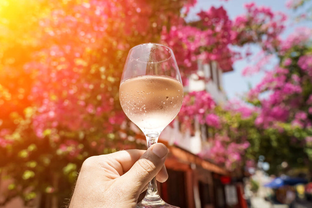 Glass of wine in hand. A glass of young fresh rose wine against the backdrop of a summer cafe in a Mediterranean seaside tourist town in the summer under sunlight. Summer, travel, lifestyle - Photo, Image