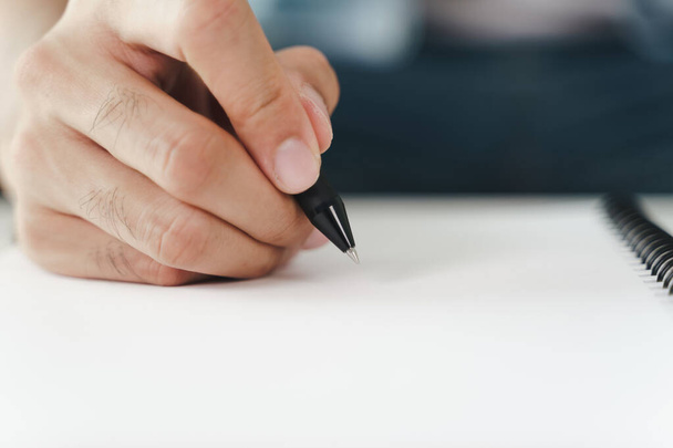 Gros plan de jeune homme en tissu décontracté mains écriture sur le bloc-notes, ordinateur portable à l'aide d'un stylo bille sur la table. - Photo, image
