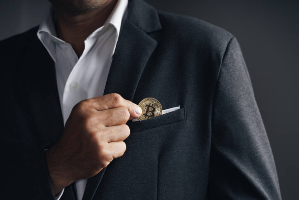 Przystojny Inwestor Businessman umieścić złoty bitcoin do kieszeni garnitur na ciemnym tle, handel, Kryptowaluta, Cyfrowa waluta wirtualna, alternatywne finanse i inwestycje Concept - Zdjęcie, obraz