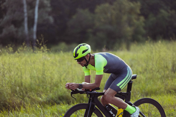 Joven triatleta masculino montando una bicicleta en el camino abierto. El deportista profesional se dedica al triatlón en el día brillante del verano - Foto, Imagen