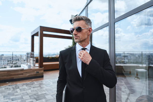 Confident mature man carrying laptop and adjusting necktie while walking on rooftop terrace - Foto, Bild