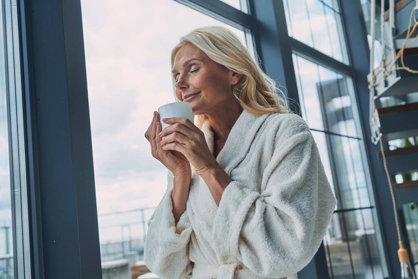 Beautiful mature woman in bathrobes drinking coffee and keeping eyes closed while standing near window at home - Фото, зображення