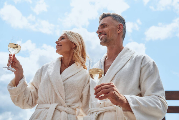 Low angle view of beautiful mature couple in bathrobes enjoying champagne while relaxing outdoors - Foto, immagini
