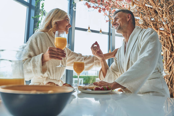 Beautiful mature couple in bathrobes enjoying breakfast together while spending time in the domestic kitchen - Foto, Bild