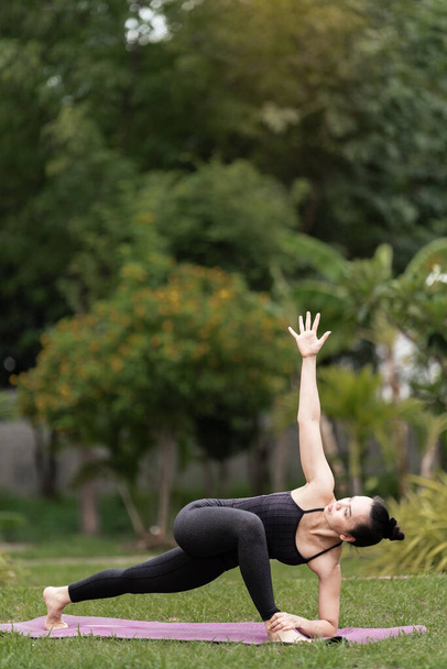 Una mujer asiática de mediana edad en traje deportivo haciendo ejercicio de yoga en la esterilla de yoga al aire libre en el patio trasero por la mañana. Mujer joven haciendo ejercicio de yoga al aire libre en la naturaleza parque público - Foto, imagen