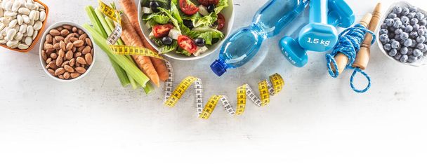 Healthy fresh salad with tomatoes surrounded with exercise equipment, carrtot celery and measuring tape - top of view. - Фото, изображение