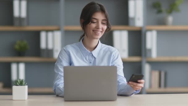 Junge Managerin tippt im Büro auf Laptop, bekommt und liest Nachricht auf Handy von geliebter Person - Filmmaterial, Video