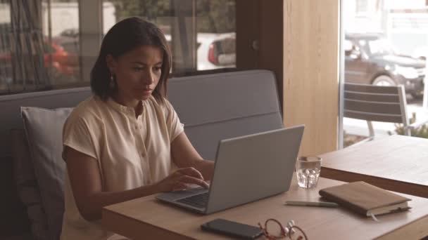 Średnie zbliżenie młodej mieszanej kobiety siedzącej przy stole w restauracji, wpisującej e-mail na przenośnym komputerze, uśmiechnięta - Materiał filmowy, wideo