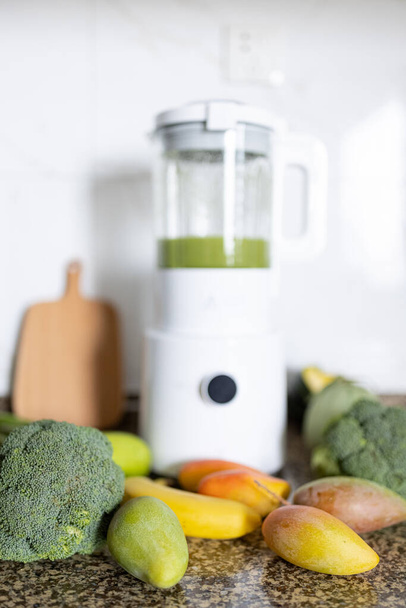 Fresh ingredients for healthy detox cocktail green color. Blender with smoothie for diet on kitchen table, next lying broccoli and other vegetables and fruits. - 写真・画像