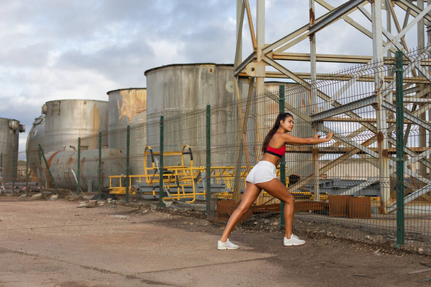 Жінка-молода спортсменка розігріває і розтягує ноги перед бігом на відкритому повітрі в промисловій зоні. Спортивний підходить жінка тренування зовні
. - Фото, зображення