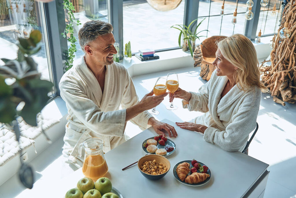 Top view of beautiful mature couple in bathrobes enjoying breakfast together while spending time in the domestic kitchen - Foto, Imagen
