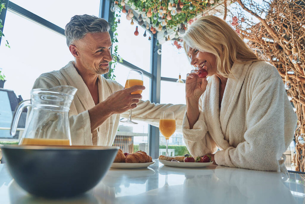 Beautiful mature couple in bathrobes enjoying breakfast together while spending time in the domestic kitchen - Photo, Image