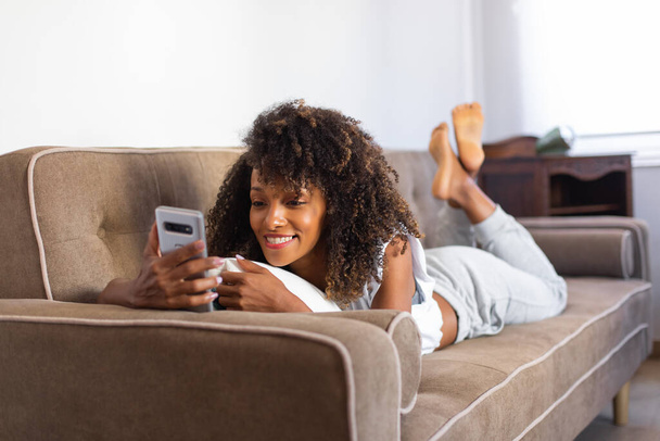 Relaxed cheerful black woman at home lying on her couch at home and using smartphone. - Photo, image