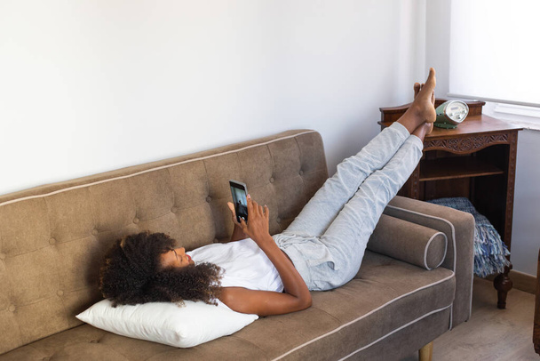 Relaxed black woman lying on her couch and using her smartphone. Relaxing, comfort and leisure at home concept. - Zdjęcie, obraz