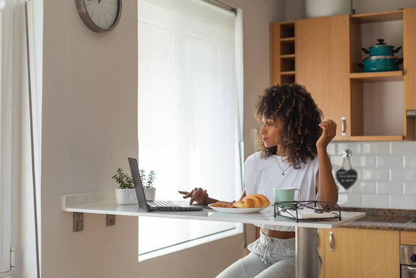 Casual comfortable young black woman taking breakfast and working from home with her laptop. - Photo, Image