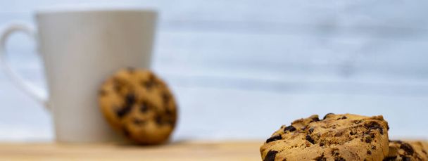 close up of cookies with chocolate pieces on a wooden table, as a background a cup for coffee or cappuccino, banner copy space area - Photo, image
