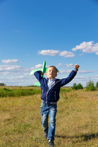 A six-year-old preschooler boy in a blue jacket launches a toy plane in a field against a blue sky with clouds on a summer day. The bright sun is shining. Scenery - Φωτογραφία, εικόνα