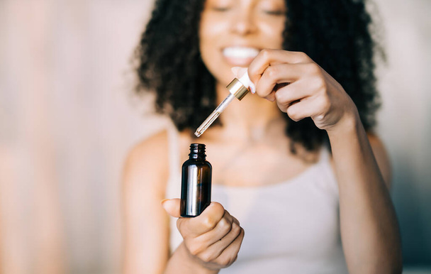 Close up african american young woman holding brown glass bottle with spa cosmeticsat hone. Beauty blogging, salon therapy, self skin care, mockup, minimalism concept - Photo, Image