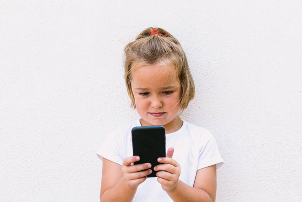 Little blond-haired girl wearing white t-shirt, looking at the mobile phone, with it turned, while taking a photo - Foto, Imagen