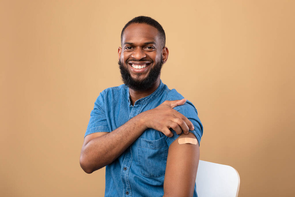 Счастливый вакцинированный черный мужчина показывает плечо с помощью браслета после прививки от коронавируса, позируя после ковидовой вакцинации - Фото, изображение