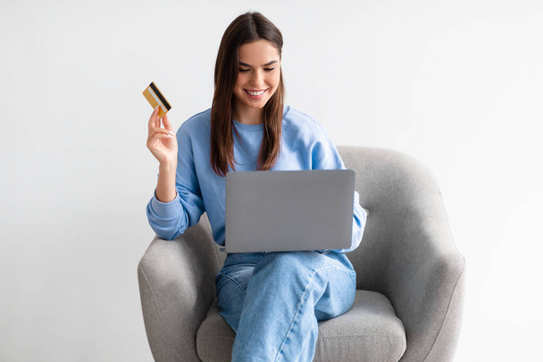 クレジットカードとノートパソコンを持つ若い女性が居心地の良いアームチェアに座って、インターネット上で商品を注文し、ウェブストアで物事を購入 - 写真・画像
