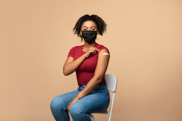 Aşılanmış yüz maskeli siyahi kadın koluna yara bandı takmış, koluna sarılmış bir şekilde oturuyor. - Fotoğraf, Görsel