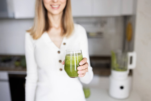 Young caucasian woman with blonde hair making detox cocktail in blender with fresh green vegetables on kitchen at home - Фото, изображение