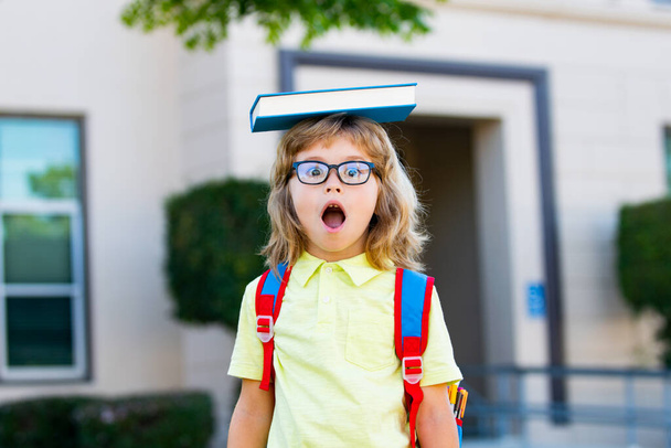 Cheerful little boy with big backpack and book having fun against school. Back to school. Concept of education and reading. - Foto, Imagen